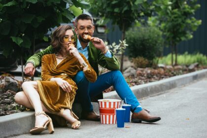 fashionable couple in velvet clothing eating fried chicken legs on street