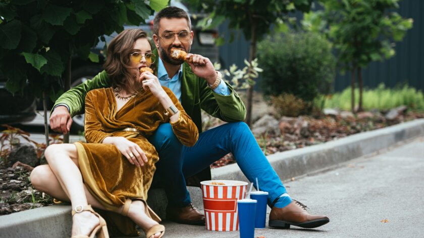 fashionable couple in velvet clothing eating fried chicken legs on street