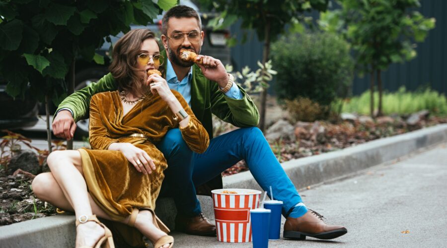 fashionable couple in velvet clothing eating fried chicken legs on street