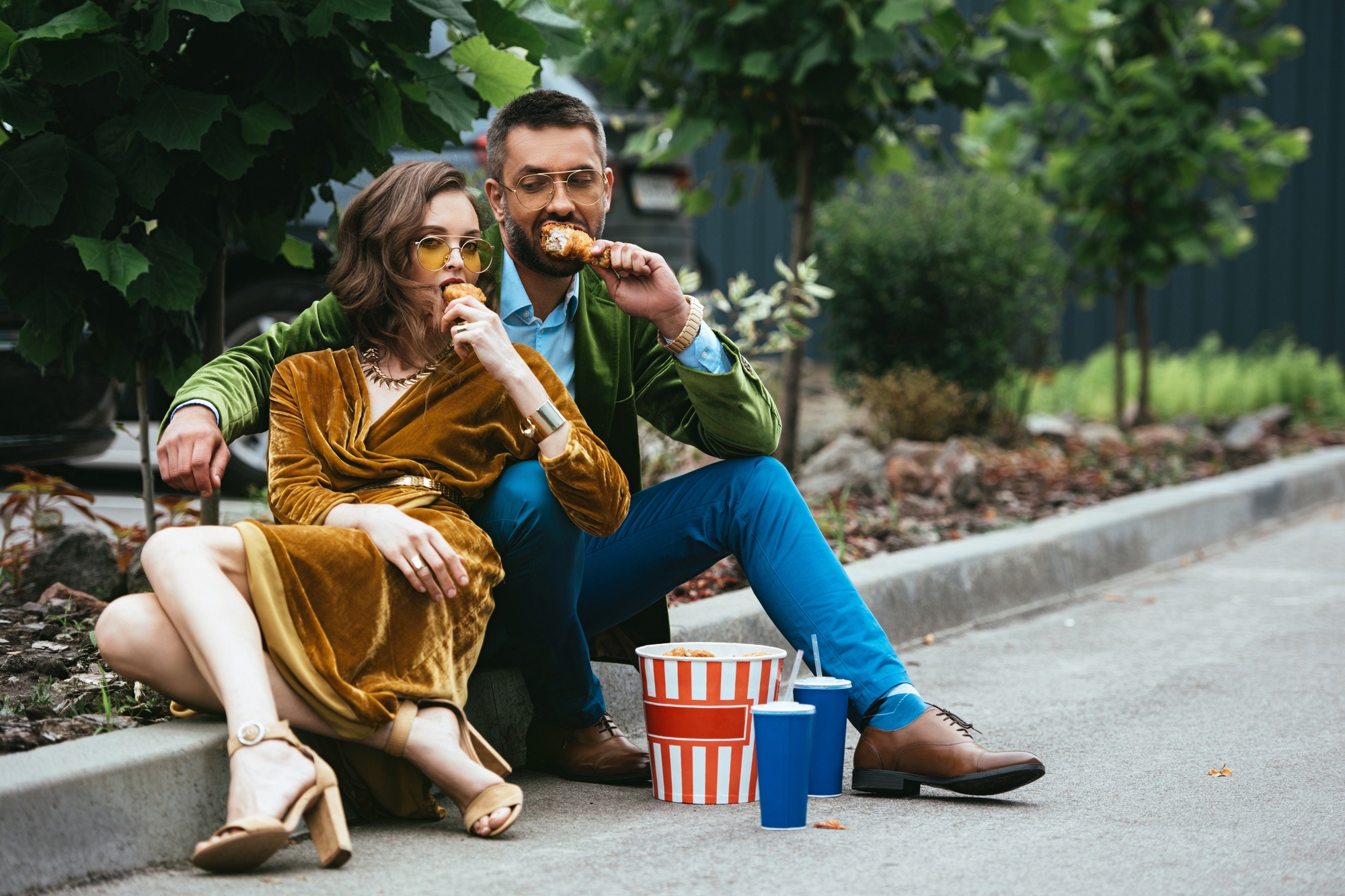 fashionable couple in velvet clothing eating fried chicken legs on street