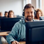 Happy mature man e-learning on desktop PC during computer class and looking at camera.