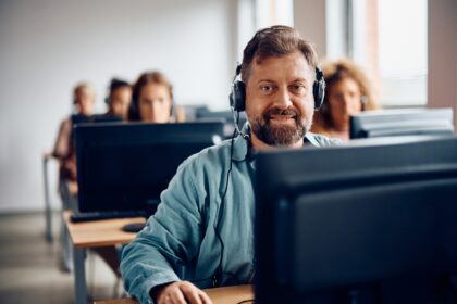 Happy mature man e-learning on desktop PC during computer class and looking at camera.
